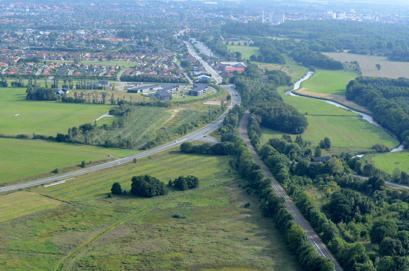 Luftfoto af et landskab med en mindre by i baggrunden og en vej og banestrækning, der løber gennem landskabet