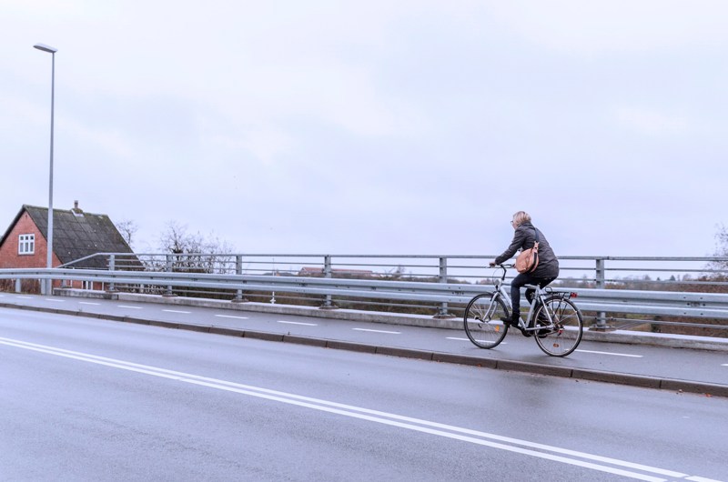 En cyklist på en bro. I baggrunden ses et rødt hus.