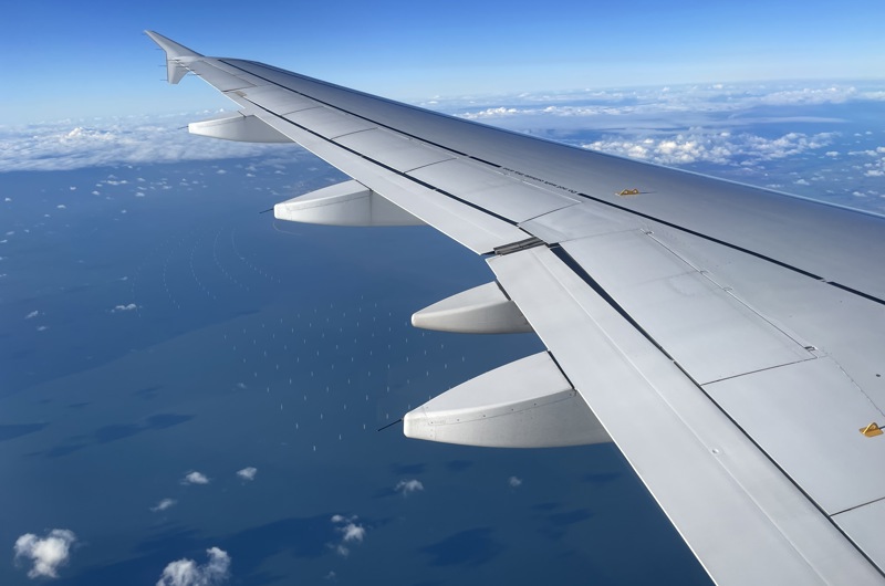Kig ud af vinduet fra et fly i luften. Udsigten viser et udsnit af flyets vinge, nogle skyer og en vindmøllepark i havet nedenunder.